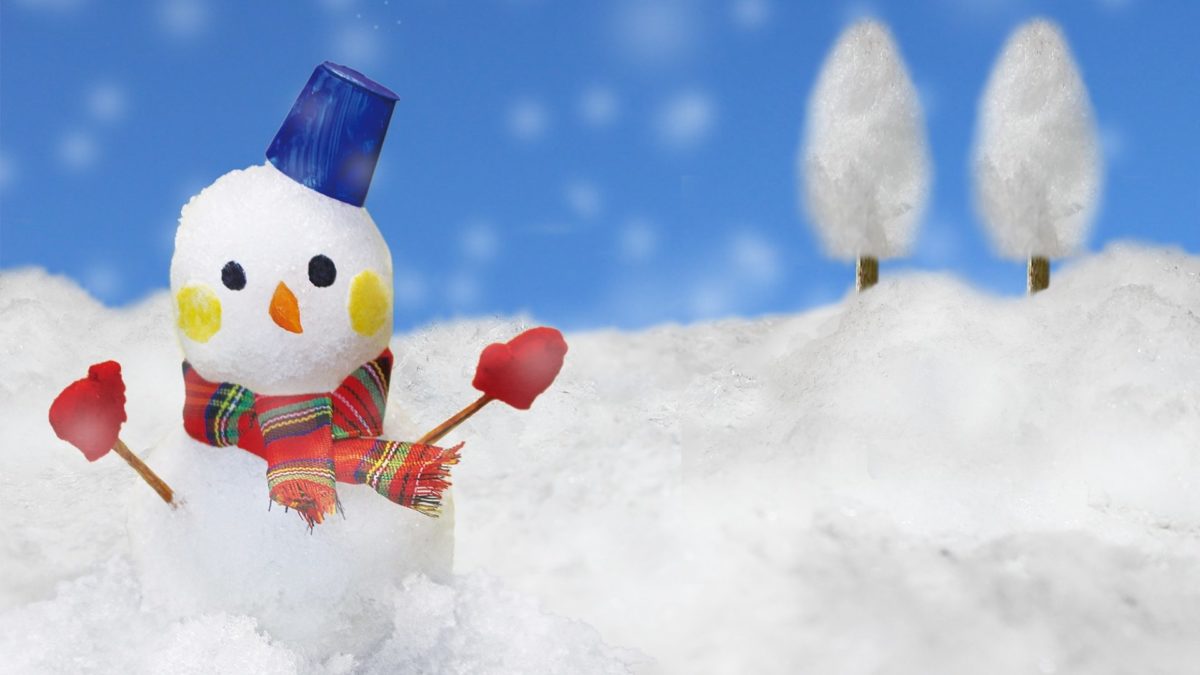Small snowman with a bucket hat, red mittens, and scarf sitting in snow under a blue sky, winter mood