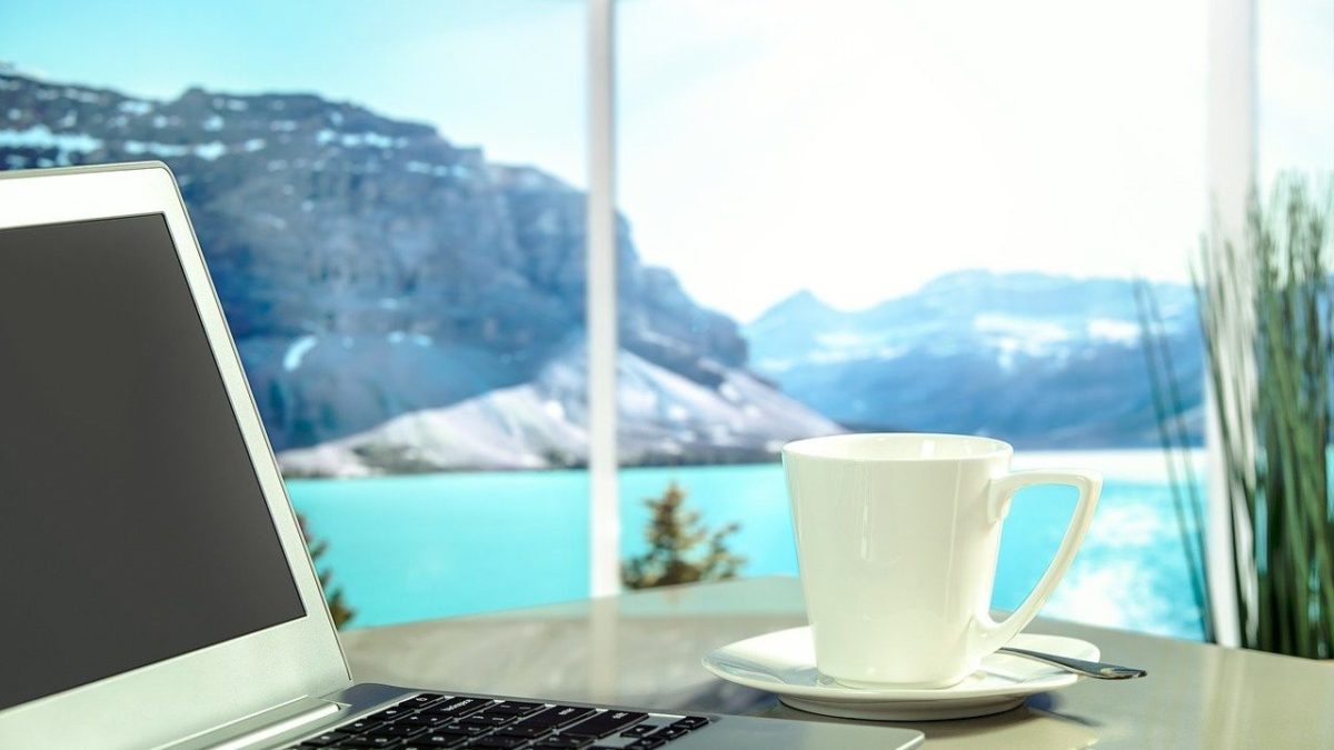 Laptop and coffee cup on a table beside a window with a scenic mountain and turquoise lake view, cool blue tones